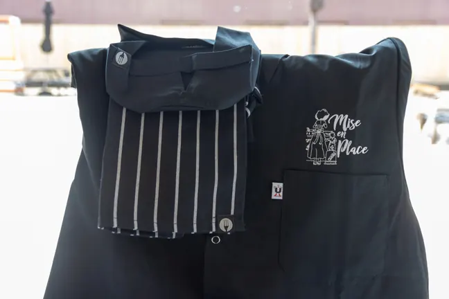 A black chef jacket and striped apron embroidered with the Mise En Place logo