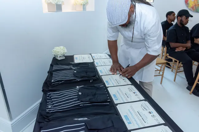A chef in whites signs certificates laid out beside folded chef jackets as students wait
