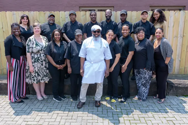 A graduating class in black uniforms with their chef instructor and supporters, posed outdoors on a brick patio