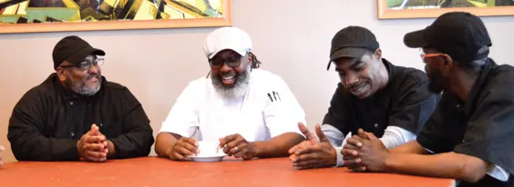 Four chefs in black and white chef coats laughing around a classroom table