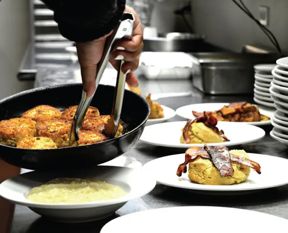 Tongs lifting golden croquettes from a skillet beside plated dishes topped with bacon