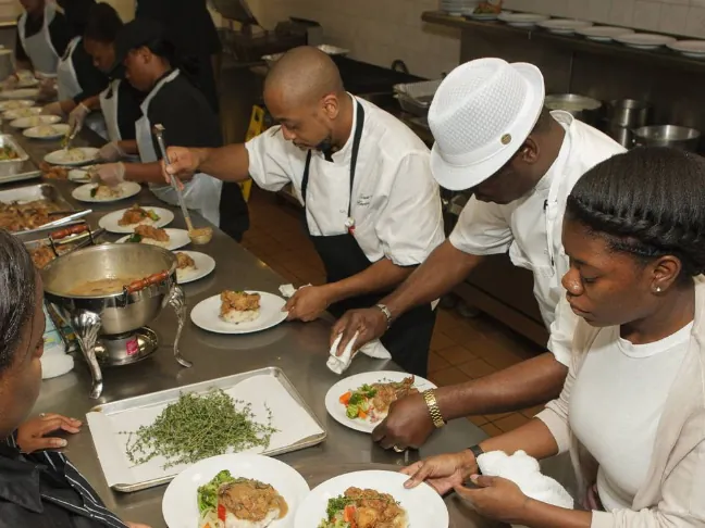 Chefs in white jackets plating dishes together at a long pass