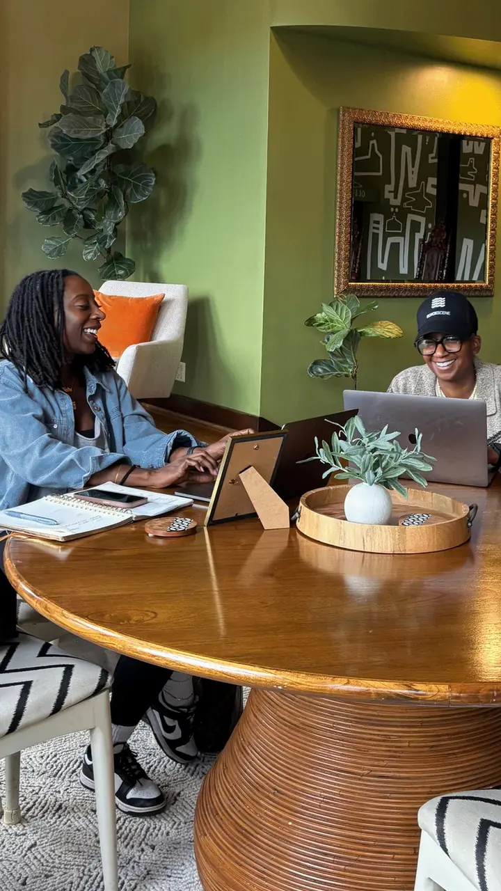 Two members working on laptops at a round wooden table in front of a sage-green wall