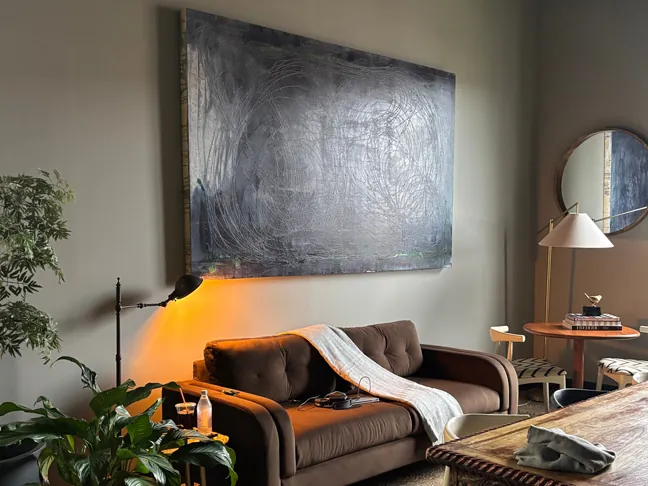 A brown leather sofa under a large abstract painting, lit by a warm lamp